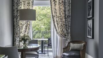 A room with a round table, a chair, a lamp, and a wicker chair. There is a balcony with a railing and a view of greenery.