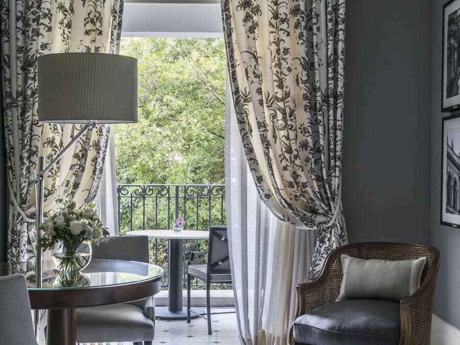 A room with a round table, a chair, a lamp, and a wicker chair. There is a balcony with a railing and a view of greenery.