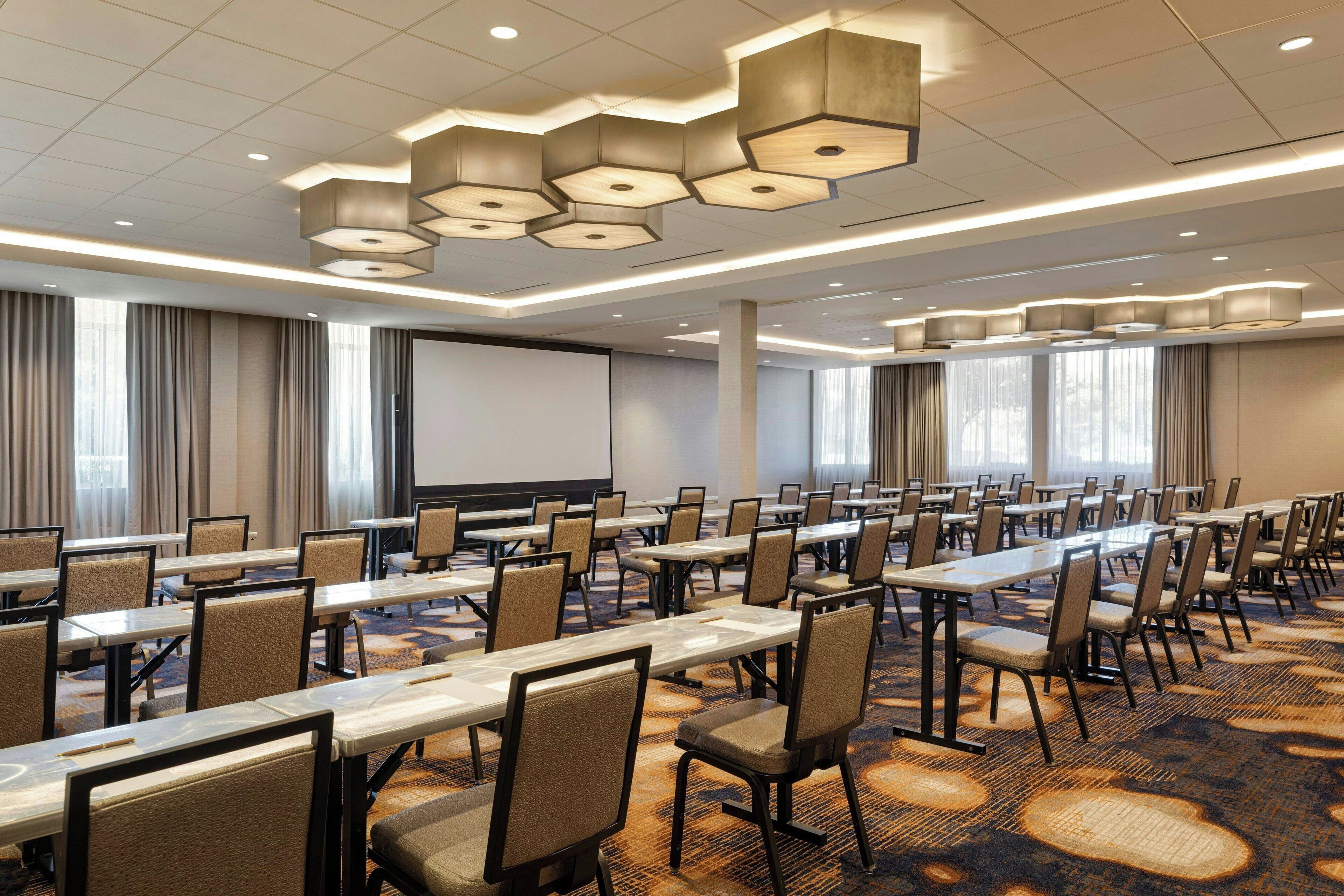 A conference room with multiple tables and chairs, a projector screen, and a large window with curtains.