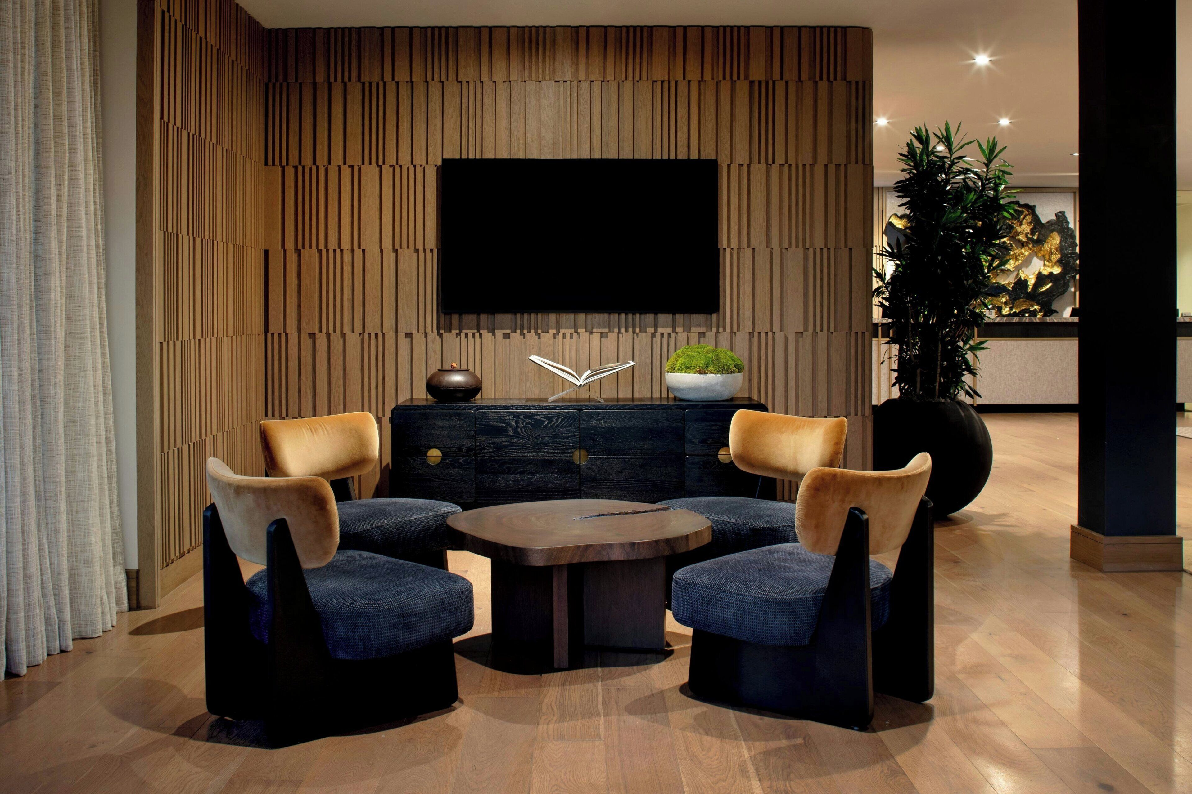 A modern living room with a round coffee table, two armchairs, a flat-screen TV mounted on a wooden wall, a dark wooden cabinet, and a large potted plant.