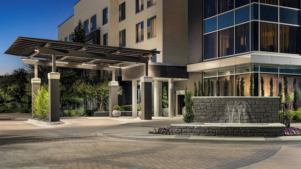 A modern hotel entrance with a covered walkway, a stone wall, and a landscaped area with plants. - The Elmore Hotel, DFW Southlake, a Tribute Portfolio Hotel (Southlake)