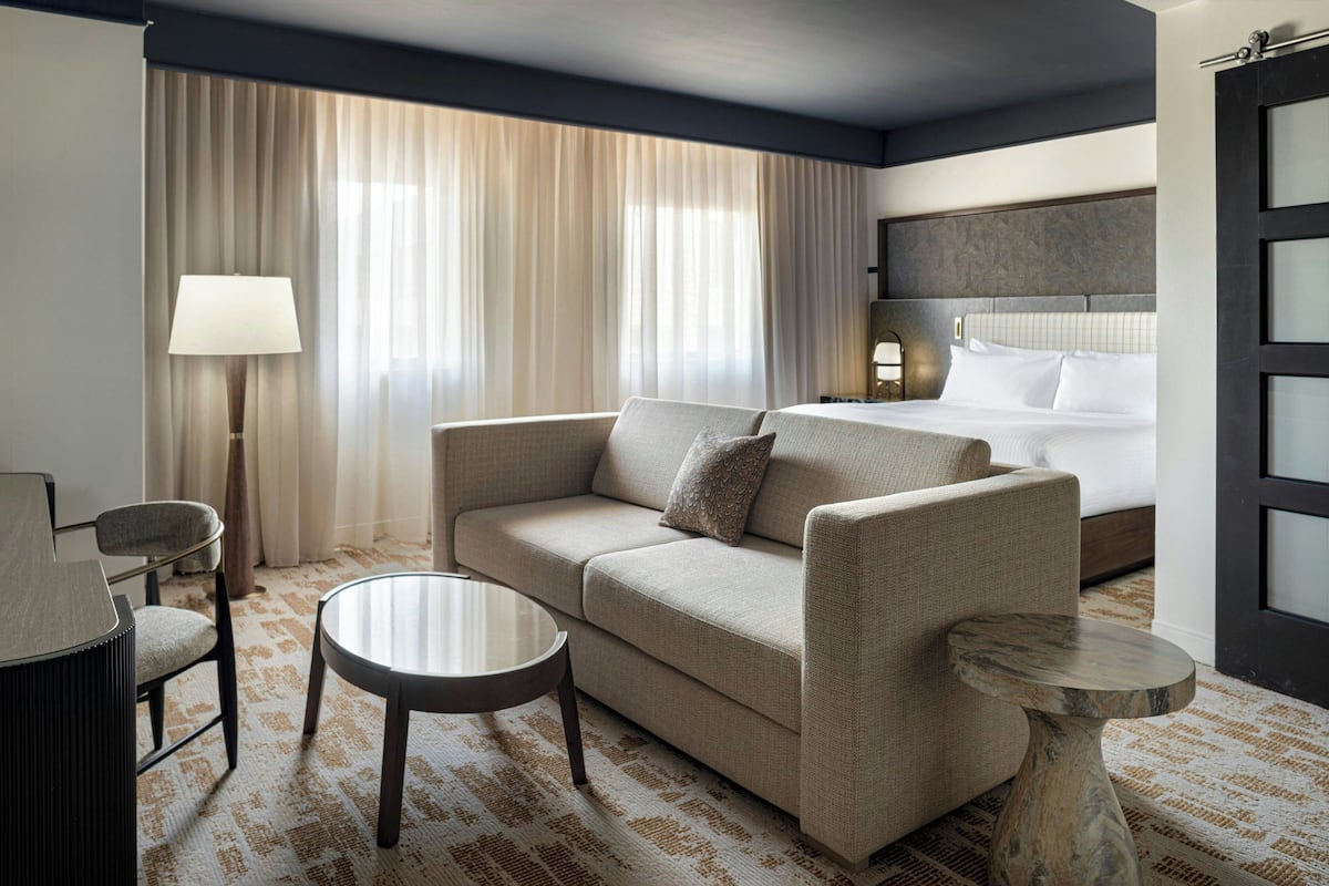 Habitación de hotel con sofá, una mesita redonda, una mesita auxiliar pequeña, una lámpara y una cama con cabecero.