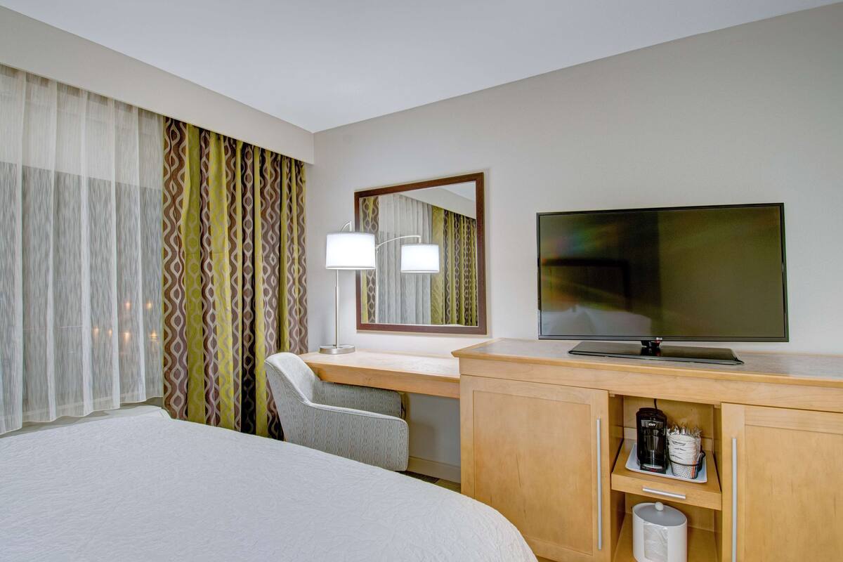 A hotel room with a bed, a desk with a television, a chair, a lamp, and a cabinet with a coffee maker.
