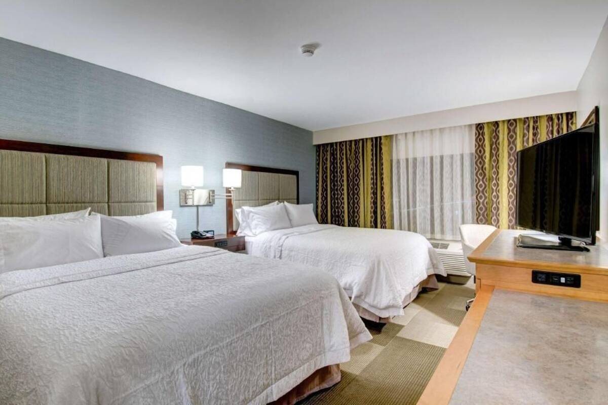 A hotel room with two beds, a flat-screen TV, a desk, and a large window with patterned curtains.