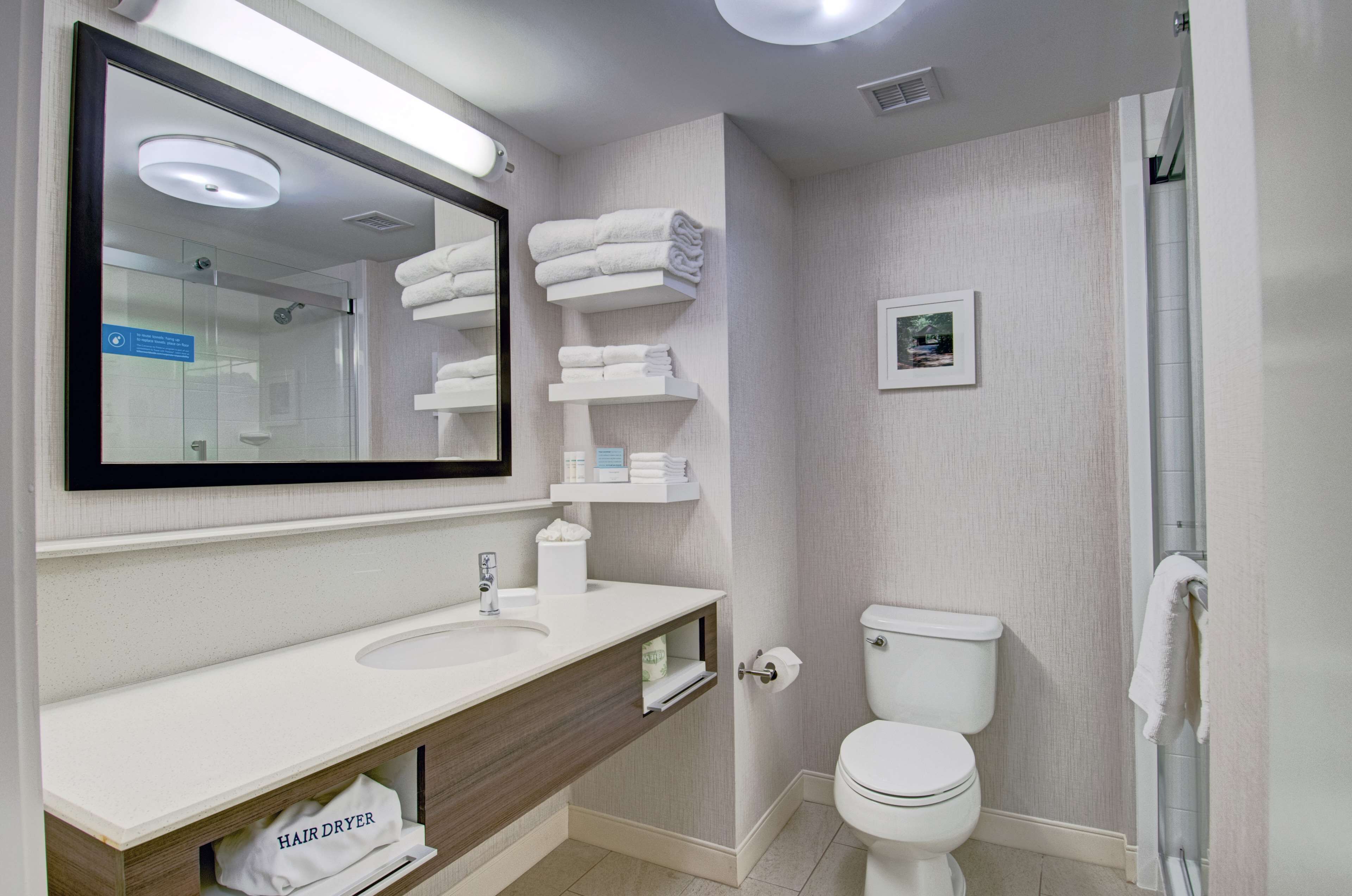 Une salle de bain moderne avec un grand miroir, un lavabo blanc et des toilettes. On y trouve des étagères avec des serviettes pliées et un sèche-cheveux posé sur le comptoir.