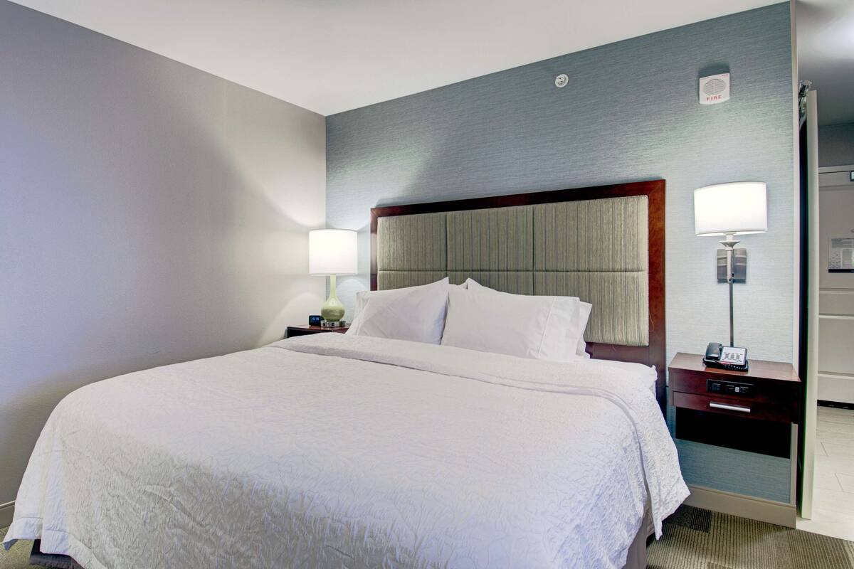 A hotel room with a large bed, a nightstand with a lamp, a bedside table with a phone, and a wall-mounted alarm clock.