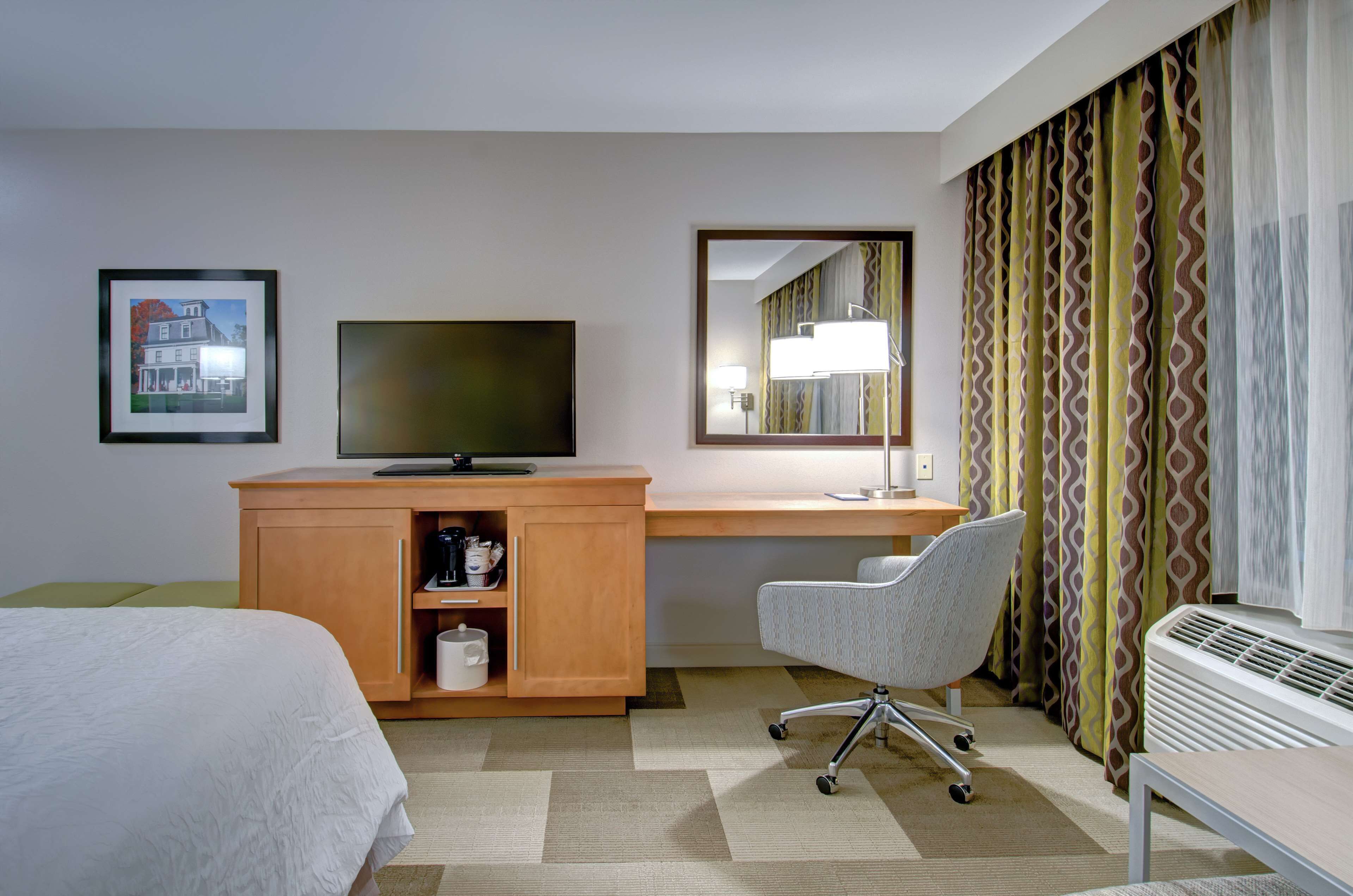 A hotel room with a bed, a desk with a computer, a chair, a TV, a mirror, and a window with patterned curtains.