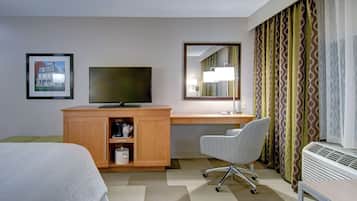 A hotel room with a bed, a desk with a computer, a chair, a TV, a mirror, and a window with patterned curtains.