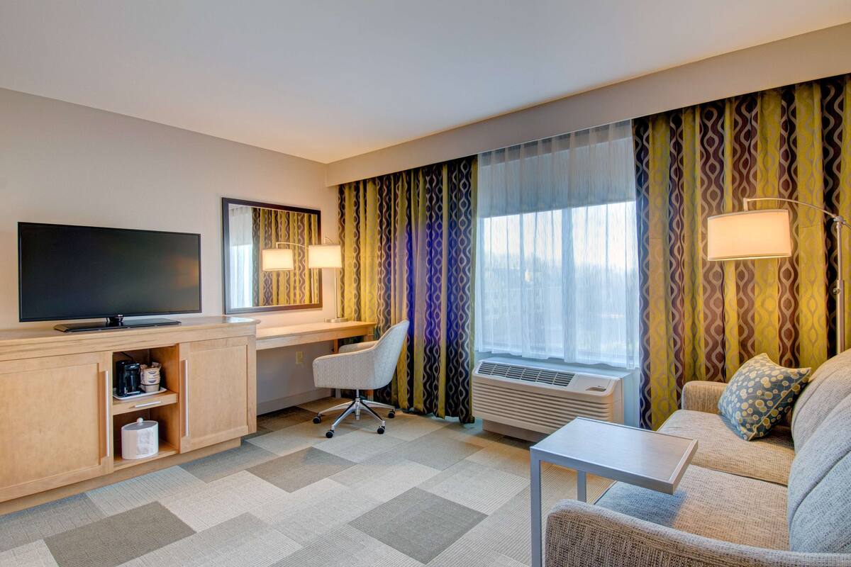 A hotel room with a flat-screen TV, a desk with a chair, a sofa, a small table, and a window with patterned curtains.