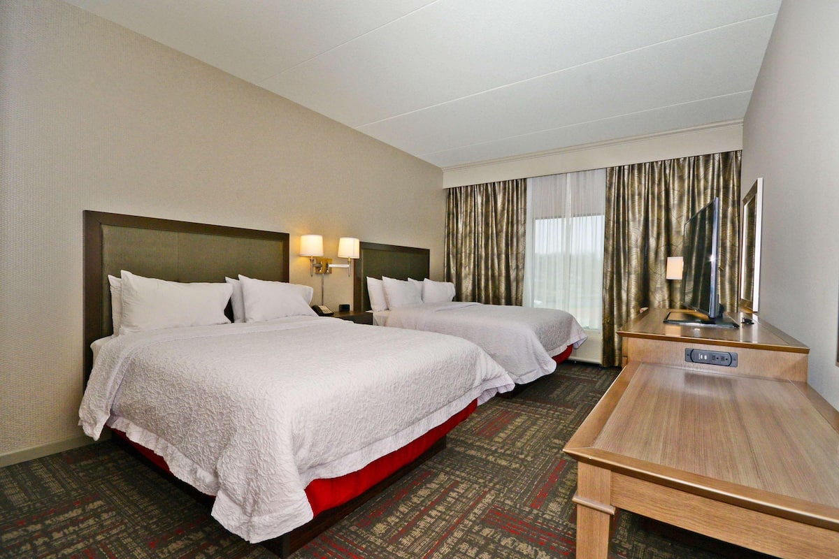 Quarto de hotel com duas camas, uma escrivaninha, uma mesa de cabeceira e uma janela grande com cortinas.