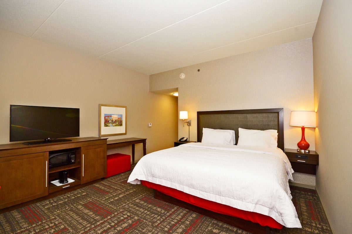 Quarto de hotel com uma cama grande, televisão de tela plana, escrivaninha com luminária e um banquinho vermelho.