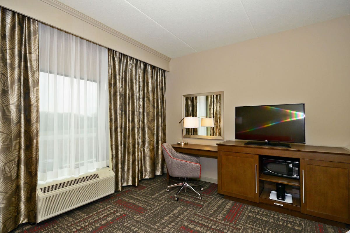 Quarto de hotel com escrivaninha, cadeira, televisão e uma grande janela com cortinas.