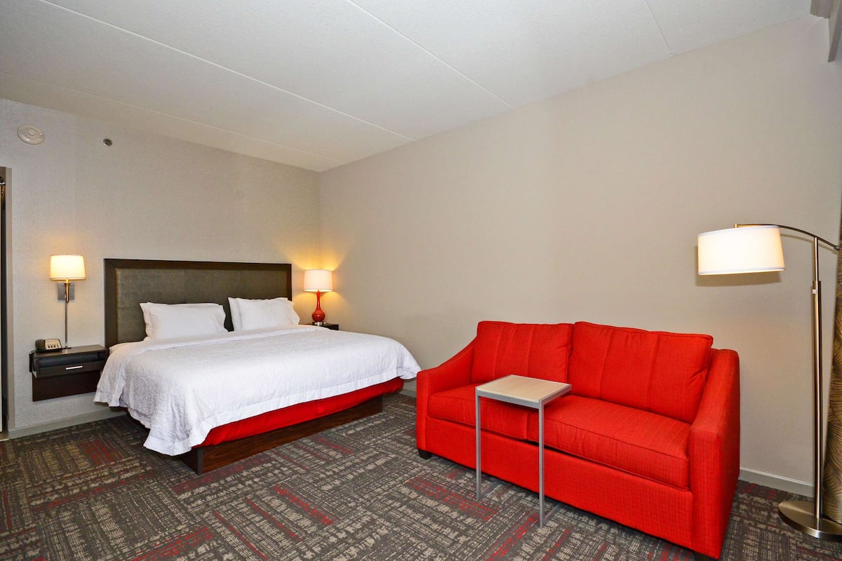 Quarto de hotel com uma cama, um sofá vermelho, uma mesinha e duas luminárias de cabeceira.