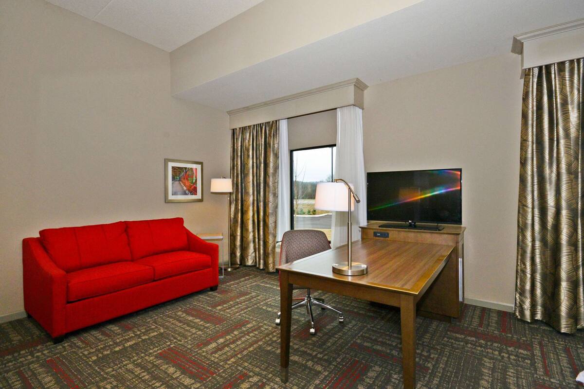 Quarto de hotel com sofá vermelho, secretária de madeira, televisão e uma janela grande com cortinas.