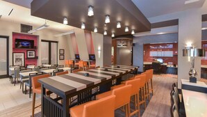 A modern restaurant with long communal tables and orange chairs, a flat-screen TV, and a chandelier. - Hampton Inn Lumberton (Lumberton)