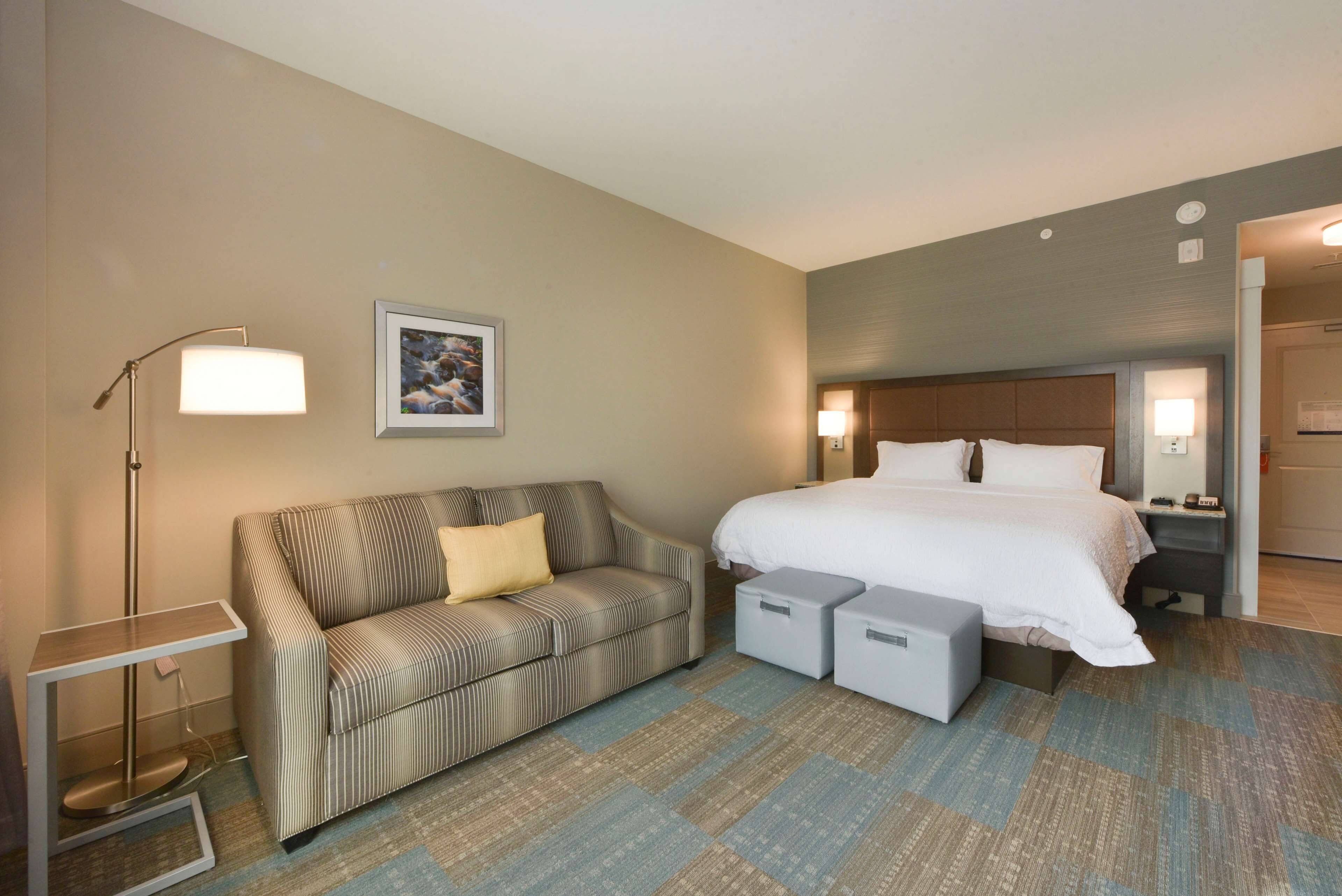 Quarto de hotel com cama, sofá, mesa de cabeceira, abajur e um quadro na parede.