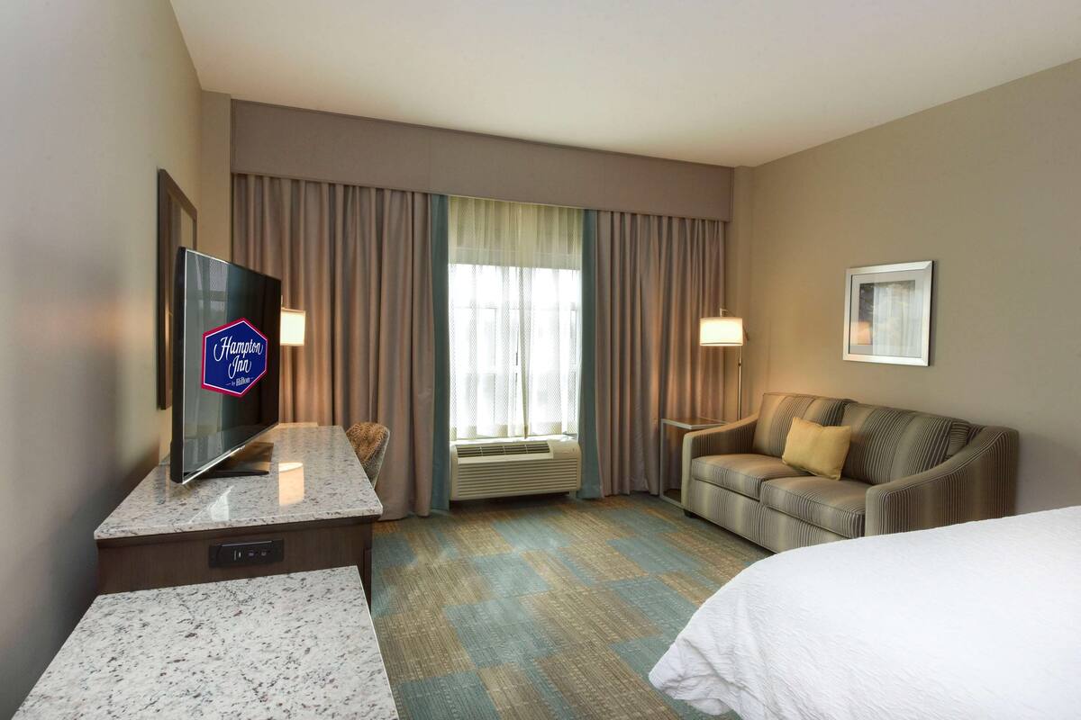 A hotel room with a flat-screen TV, a sofa, a bed, and a large window with curtains.