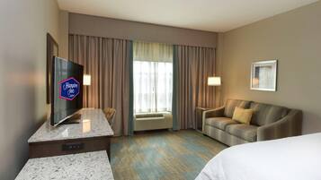 A hotel room with a flat-screen TV, a sofa, a bed, and a large window with curtains.