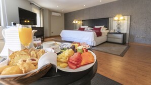 Un bufé de desayuno con diversos pasteles, frutas y un vaso de zumo de naranja sobre una mesa en una habitación de hotel con una cama y un televisor al fondo.