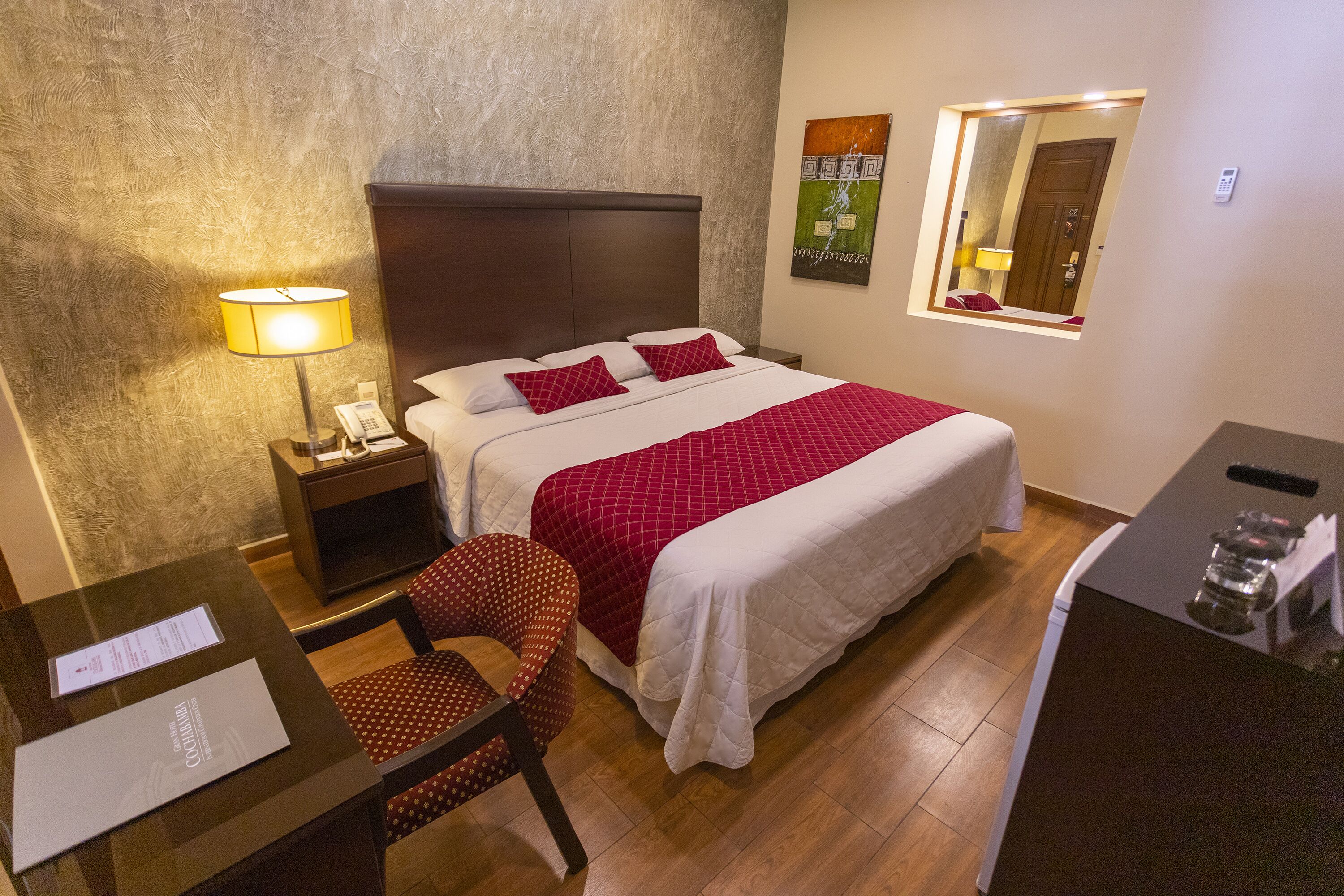 A hotel room with a bed, a desk with a lamp, a chair, and a small table with a phone.
