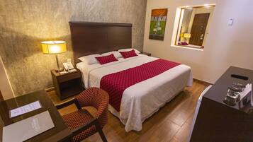 A hotel room with a bed, a desk with a lamp, a chair, and a small table with a phone.