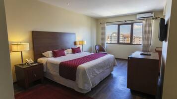 A hotel room with a bed, bedside lamps, a desk, a chair, and a window with a city view.