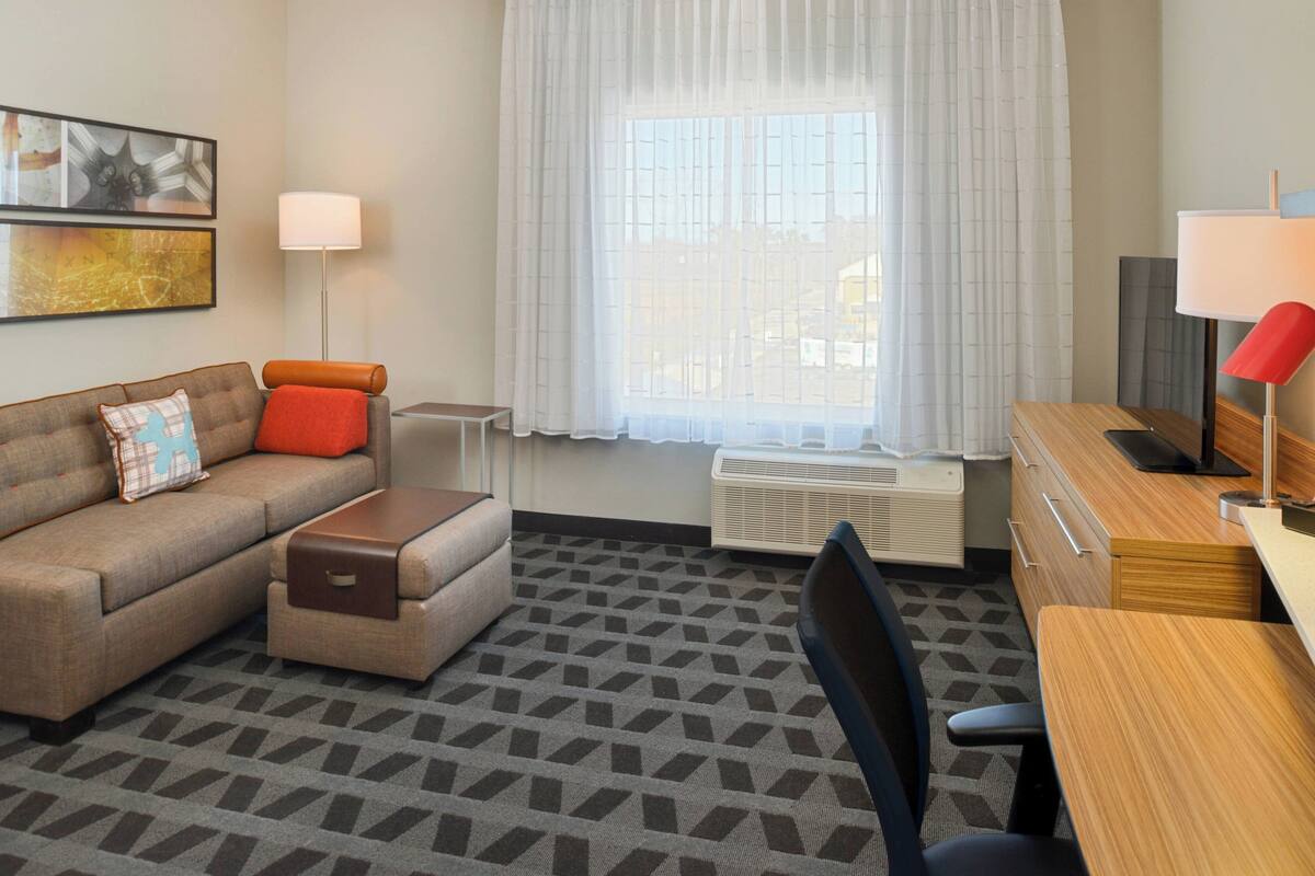 Ein Hotelzimmer mit Sofa, Schreibtisch, Stuhl, Lampe, einem fenster mit Vorhängen und einer Heizung.