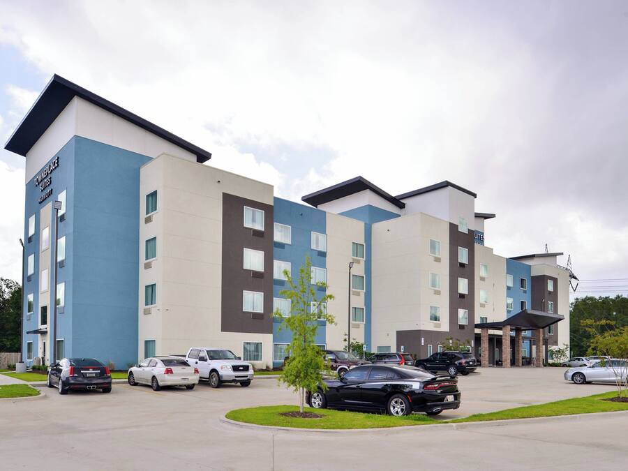 A multi-story hotel with a modern design, featuring a parking lot with several cars and a small landscaped area with a tree.