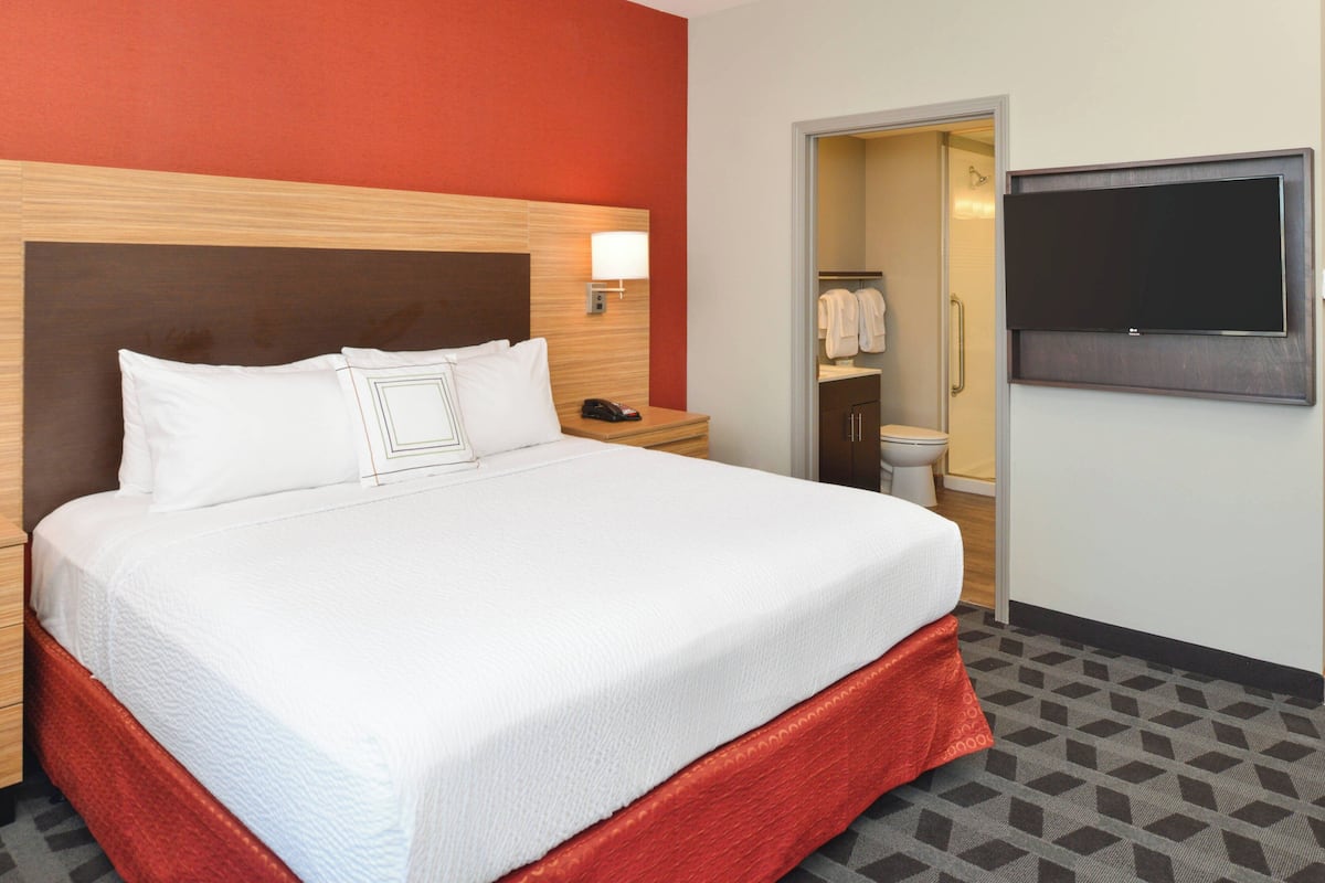 Ein Hotelzimmer mit einem großen Bett, einem Nachttisch mit Lampe, einem an der Wand montierten Fernseher und einem durchgehenden Türspalt sichtbaren Badezimmer.