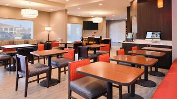 A modern hotel lobby with multiple seating areas, including tables and chairs, a reception desk, and a large TV screen.