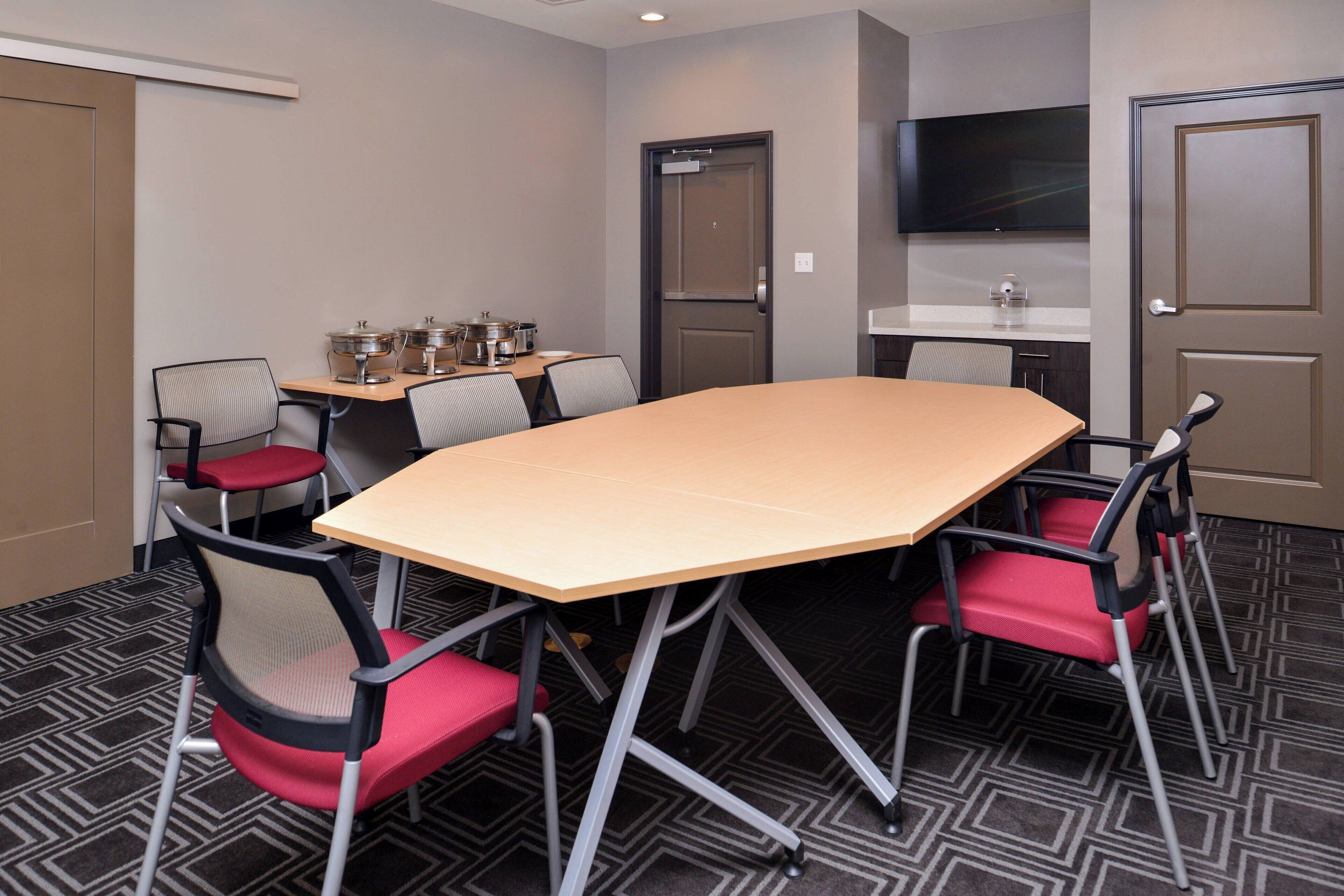A conference room with a large table, four chairs, a television, and a kitchenette area.