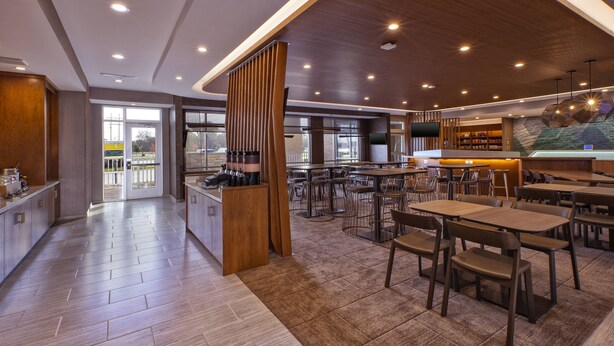 A modern restaurant with a wooden bar, multiple tables and chairs, and a large window providing a view of the outside. - SpringHill Suites by Marriott St. Joseph Benton Harbor (Benton Harbor)