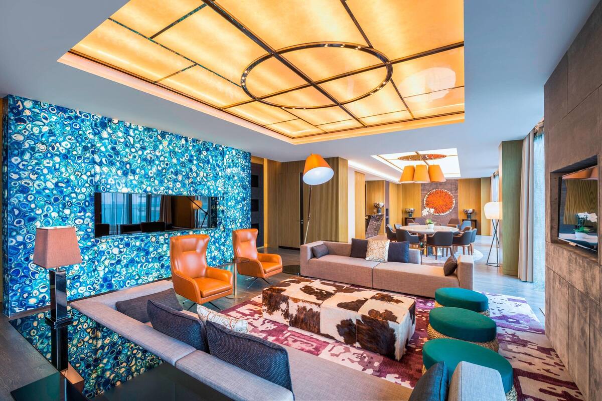 A modern living room with a blue patterned wall, orange chairs, a sofa, and a dining area with a round table.
