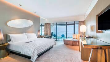 A modern hotel room with a large bed, a desk, a flat-screen TV, and a view of the sea.