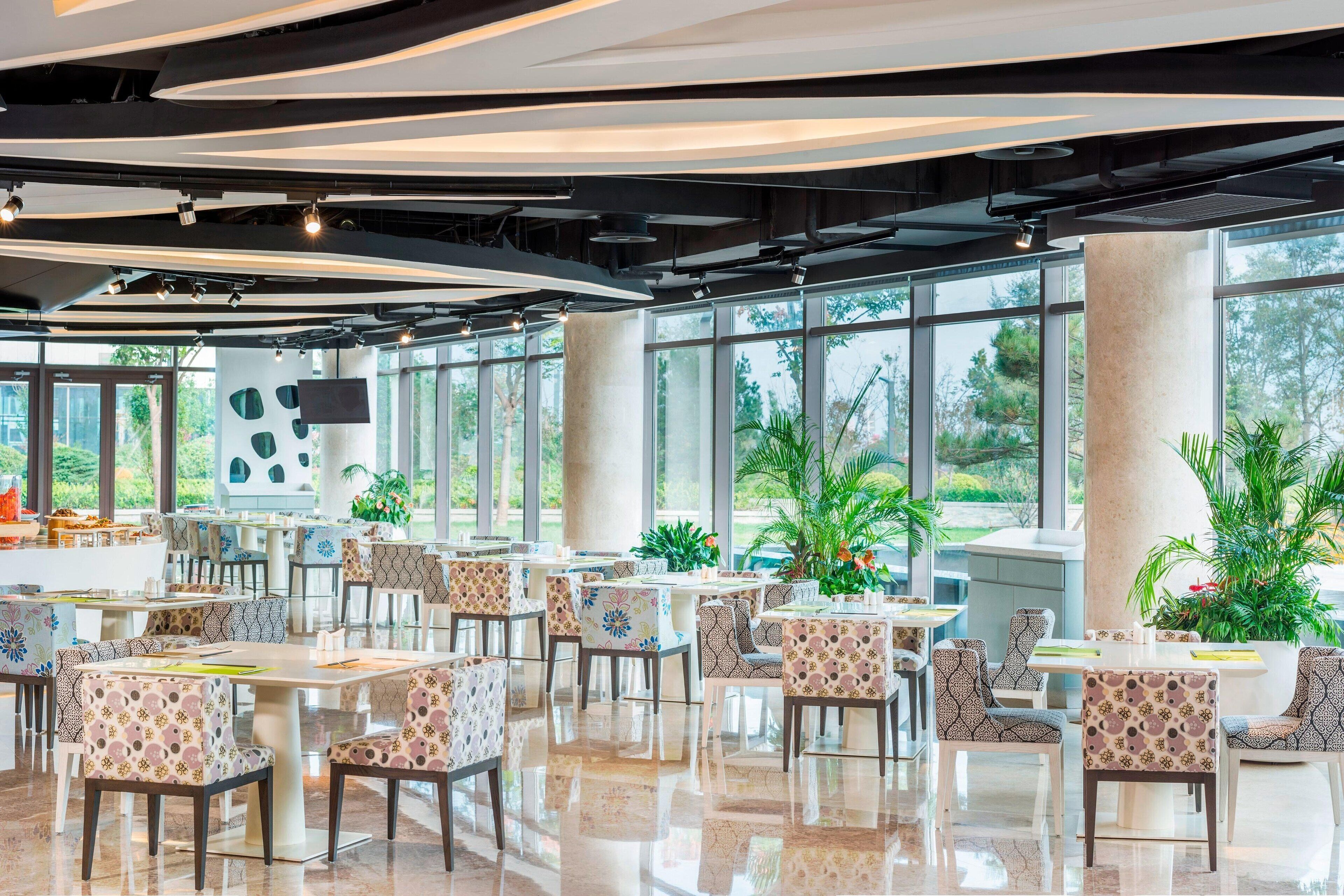 A spacious indoor dining area with tables and chairs, large windows, and potted plants.