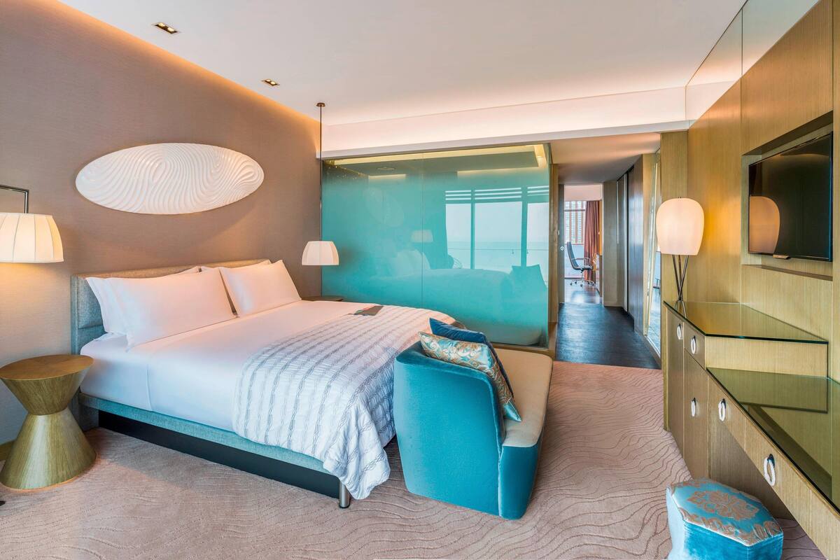 A modern hotel room with a large bed, a seating area, a TV, and a view of the city.