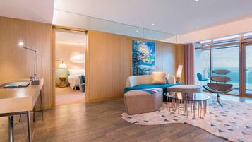 A modern hotel room with a wooden floor, a desk with a lamp, a sofa with a colorful throw, a round table with chairs, and a large window with a view.