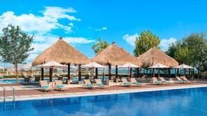Una piscina con sillas de descanso y sombrillas de paja, rodeada de árboles y un cielo despejado.