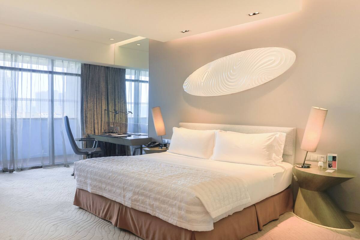 A hotel room with a large bed, a desk, a chair, and a view of the city through the window.