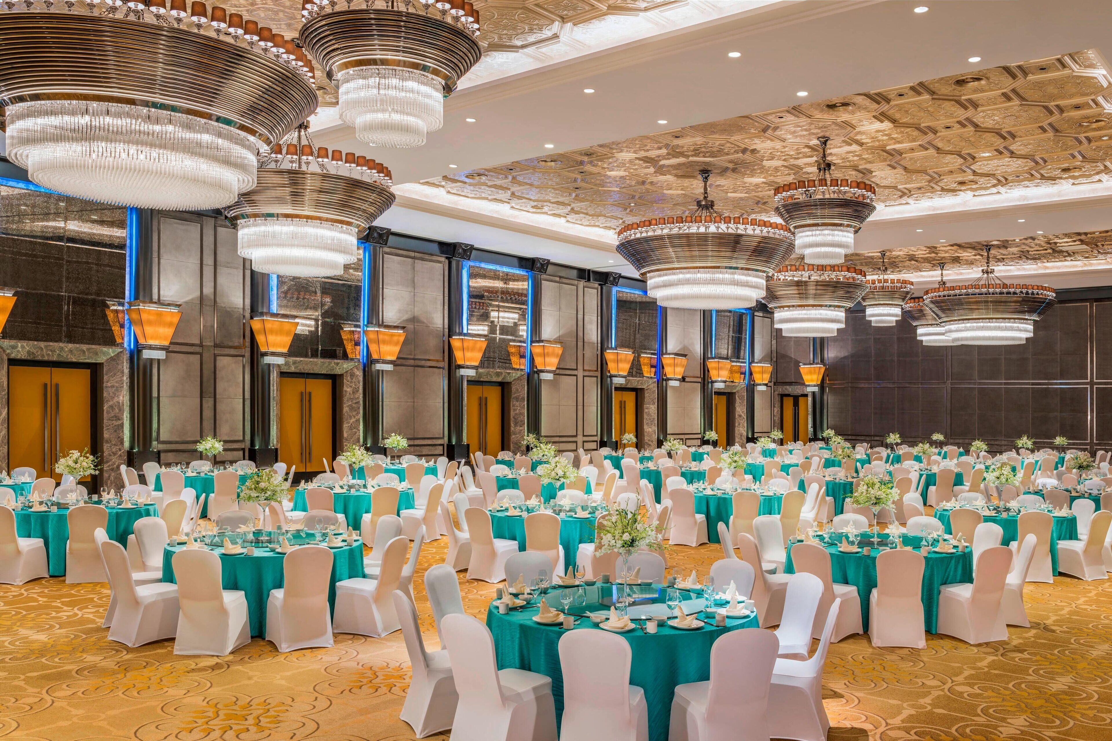 A banquet hall with round tables, white chairs, teal tablecloths, and large chandeliers.