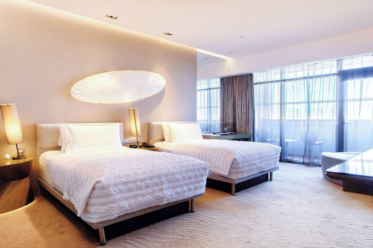 A hotel room with two beds, a large window with curtains, a modern lamp, and a small table.