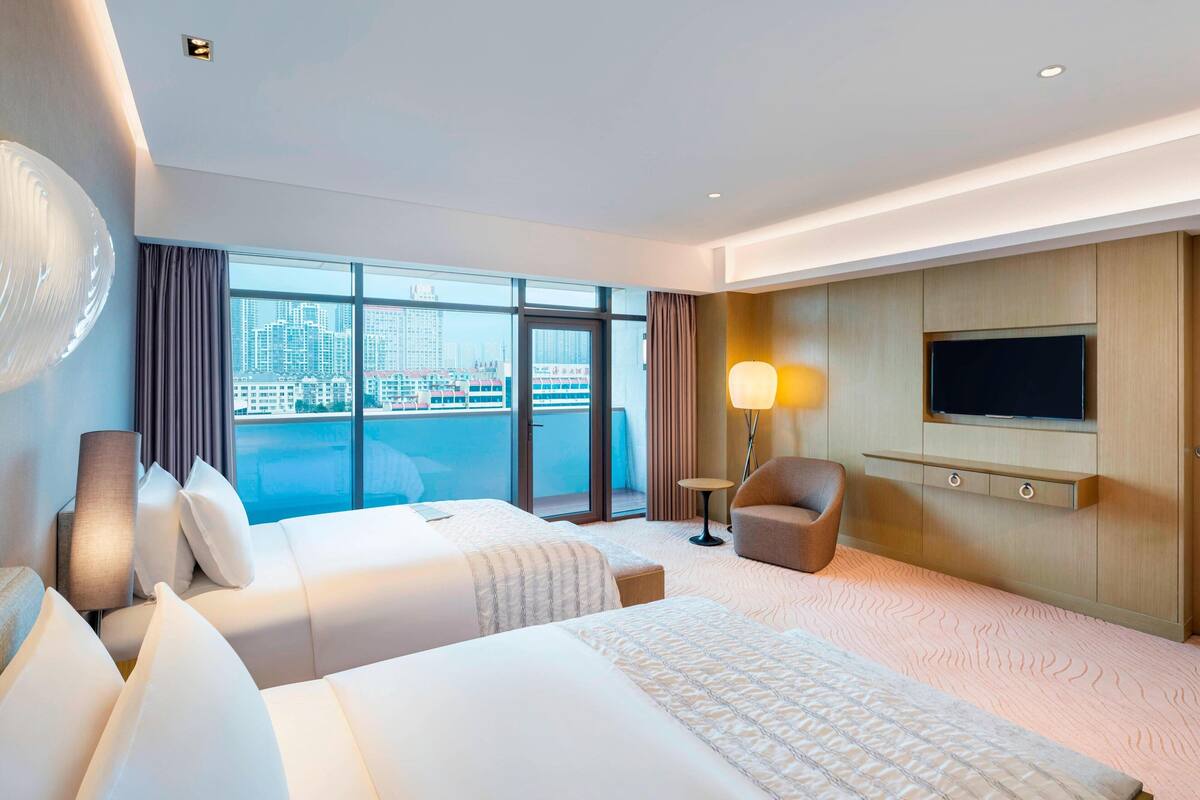 A hotel room with two beds, a TV, a chair, and a view of the city.