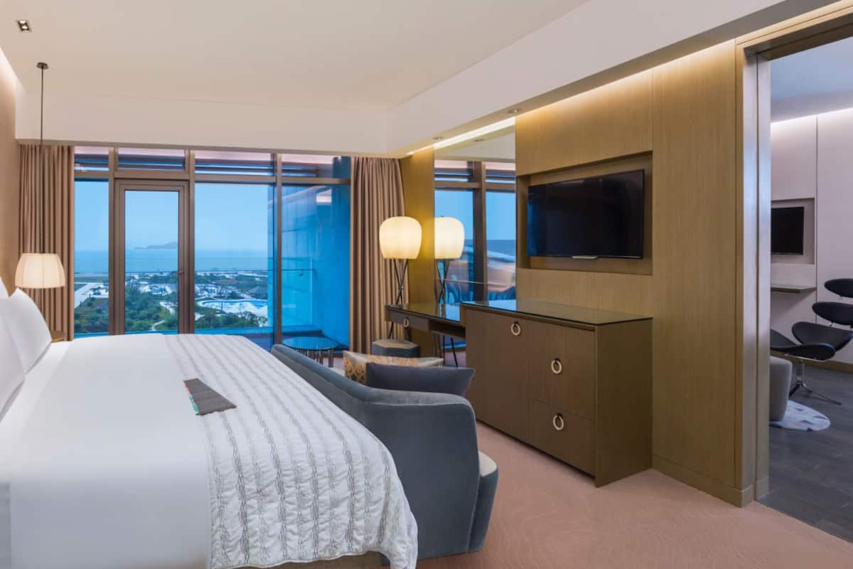 A modern hotel room with a large bed, a desk, a TV, and a view of the city.