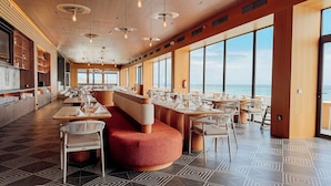 A modern restaurant with a long counter, high ceilings, and large windows overlooking the sea.