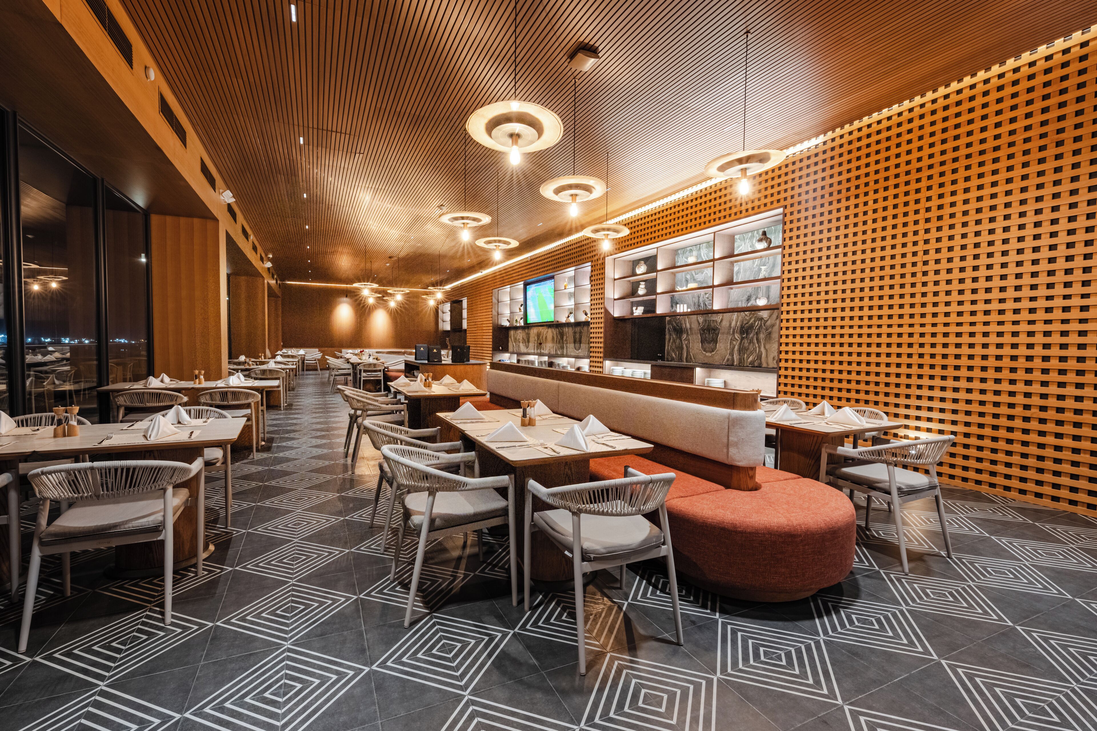 A modern restaurant interior with geometric patterned flooring, wooden ceiling panels, and a central seating area with a red ottoman.