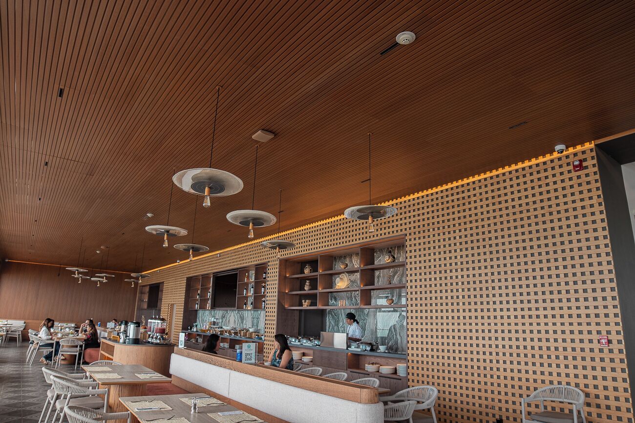 A modern restaurant with wooden ceiling and walls, a counter with a display of dishes, and seating areas with tables and chairs.