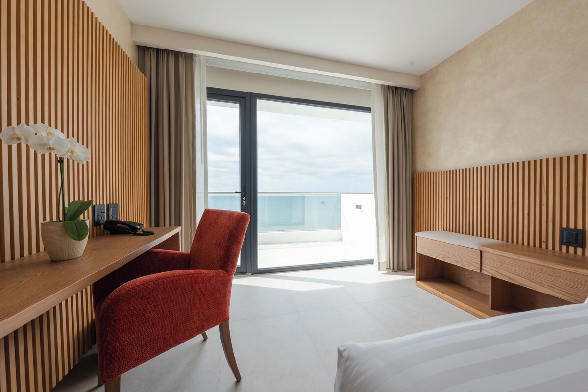 A hotel room with a wooden desk, a red chair, a bed, and a large window offering a view of the sea.