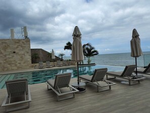 Zona junto a la piscina con sillones de descanso y sombrillas, con vistas a una extensión de agua.