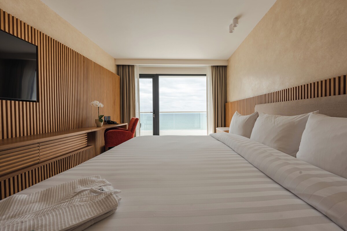 Habitación de hotel con una cama grande, un televisor montado en la pared, un escritorio con silla y vistas al cielo a través de un ventanal amplio.