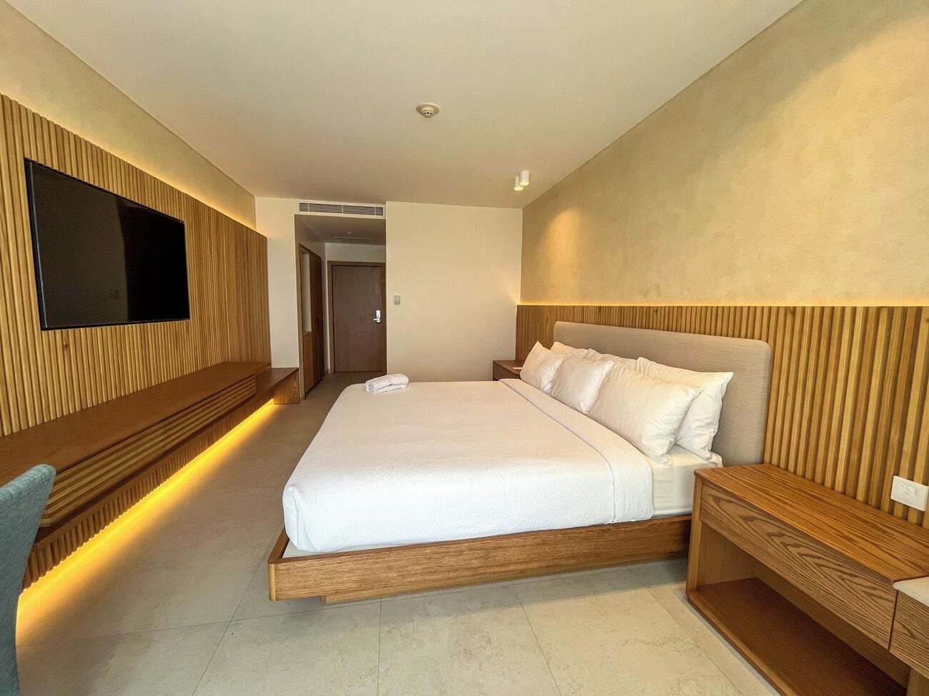 A modern hotel room with a large bed, a flat-screen TV mounted on the wall, and a wooden headboard.